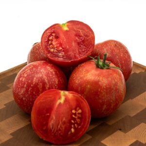 Several Justin’s Fire Opal tomatoes, red and speckled, are arranged on a wooden cutting board; two are sliced in half to show their juicy interior and seeds.